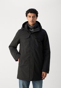 Parka de invierno negra con capucha, cierre de cremallera, dos bolsillos en el pecho y una bufanda de punto. La tela parece ser resistente al agua y texturizada.