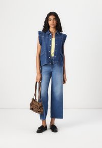 Denim ermløs jakke med volanger på skuldrene, båret over en lys gul topp. Kombinert med vide blå jeans og en brun håndveske. Sorte sko.