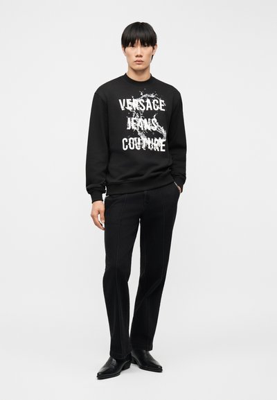 Homme debout portant un sweat-shirt noir avec le texte blanc "Versace Jeans Couture" et un pantalon noir avec des chaussures noires sur un fond uni.