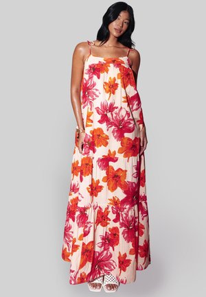 Vrouw met mouwloze, enkellange jurk met rood en oranje bloemenpatroon en witte open-toe sandalen, staand tegen een grijze achtergrond.