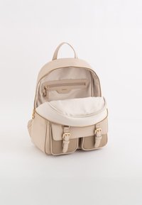 Mochila de imitación de cuero beige con herrajes dorados, que cuenta con un bolsillo con cremallera y dos compartimentos frontales, forro suave y forma redondeada.