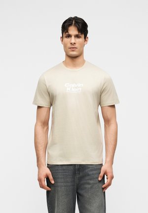 Férfi bézs színű Calvin Klein pólóban és sötétszürke farmerban, előre nézve, tiszta fehér háttér előtt.