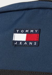 Tamno plavi materijal s pravokutnom izvezenom značkom koja prikazuje "TOMMY" iznad "JEANS" i pravokutni logotip u crvenoj, bijeloj i plavoj boji.