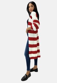 Długi kardigan w czerwono-białe paski z prążkowaną fakturą, otwarty z przodu, z opuszczonymi rękawami na ramionach, zestawiony z dopasowanymi dżinsami i czarnymi loafersami.