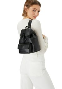 Zaino in pelle nera con alette ripiegabili, tasca frontale, spallacci regolabili e una superficie liscia, indossato da una persona in abbigliamento bianco.