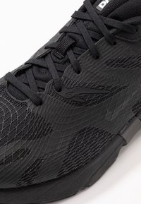 Black athletic shoe featuring textured synthetic upper, dynamic grooves, and black laces, with a reinforced sole for durability.
