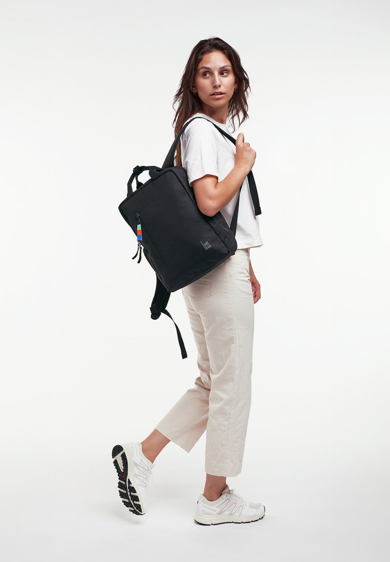 Femme en t-shirt blanc, pantalon beige et baskets blanches, marchant avec un sac à dos noir sur une épaule, regardant vers sa droite.