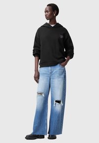 Felpa nera con cappuccio e logo, abbinata a jeans a gamba larga azzurri chiari con dettagli strappati. Stivali neri chunky completano il look.