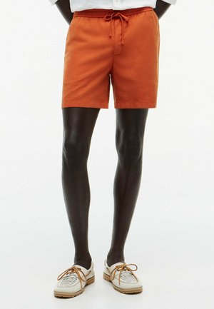 Osoba nosząca pomarańczowe szorty z troczkiem i białe plecione skórzane buty z brązowymi sznurowadłami stoi na białym tle.