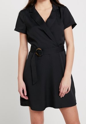 Kobieta ubrana w czarną sukienkę kopertową z krótkim rękawem, w pasie z paskiem i okrągłą klamrą, stojąca na jednolitym tle.