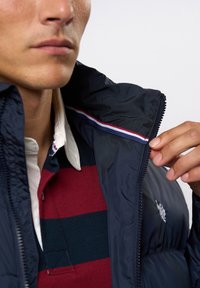 Marineblauwe gewatteerde jas met een rits, met een gestreepte accent aan de kraag en een geborduurd logo op de borst. Strepen in rood en wit.