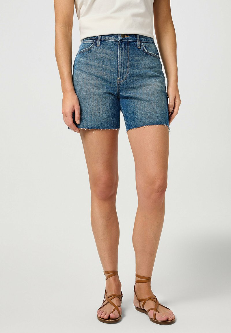 Farkkushortseissa on keskisininen pesu, korkea vyötärö, revitty helma ja klassinen viiden taskun muotoilu. Yhdistetty ruskeiden hihnasaappaiden kanssa.