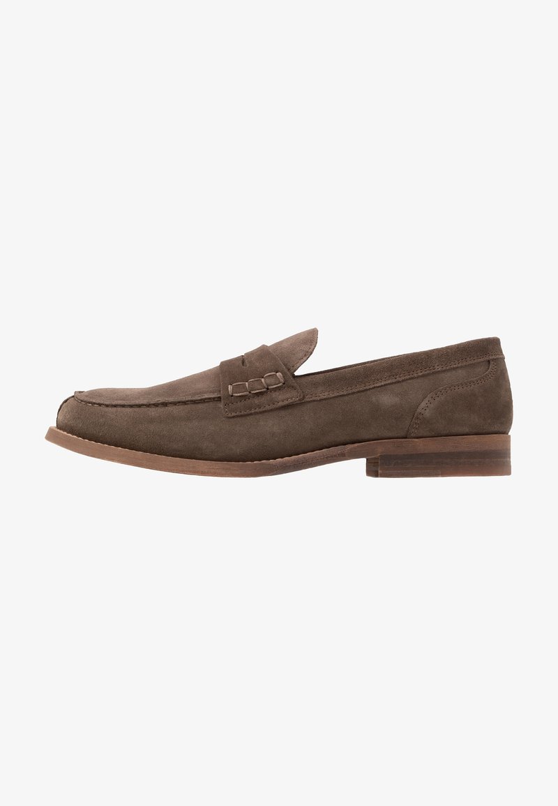 Mocassin en suede marron avec un bout arrondi, un talon plat et une sangle décorative sur le dessus. Texture lisse avec des détails de couture simples.