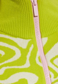 Grüner und weißer gemusterter Pullover mit hochgeripptem Kragen, ausgestattet mit einem pinken Reißverschluss in der Mitte. Weicher Stoff, kräftiger Farbkontrast.