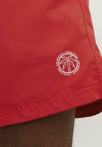 Rode zwemshorts van lichtgewicht stof hebben een palmboomlogo in het wit op het onderbeen en contrasterende stiknaden.