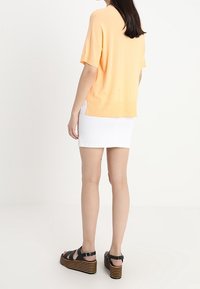 Vrouw die staat, gekleed in een perzikkleurig shirt met korte mouwen, witte minirok en zwarte platformsandalen met gestreepte zolen, met de rug naar de camera op een witte achtergrond.