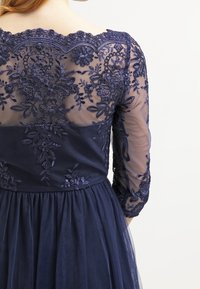Granatowa sukienka z przezroczystymi długimi rękawami, z misternym kwiatowym haftem na plecach oraz dopasowaną talią prowadzącą do zwiewnej spódnicy.
