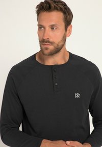 Chemise noire à manches longues avec un col rond, patte de boutonnage à trois boutons et logo blanc sur la poitrine gauche ; tissu en coton doux, poignets côtelés.