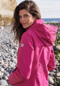 Fuchsia lightweight jacket with a hood, featuring stitching details and a circular logo on the left sleeve. Smooth exterior and flexible fit.