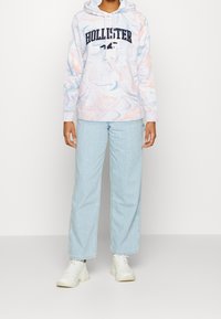 Jasno pastelowa bluza z kapturem z marmurkowymi wzorami w odcieniach niebieskiego i różowego, z kapturkiem na sznurek i logo "Hollister". Dobranie do jasnoniebieskich szerokich spodni dżinsowych i białych butów sportowych.