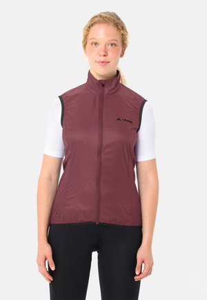 Vrouw draagt een mouwloos bordeauxrood vest met ritssluiting over een wit shirt met korte mouwen en zwarte broek, staande tegen een effen achtergrond.