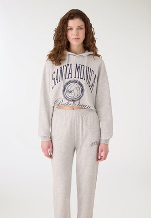 Giovane donna con capelli ricci che indossa una felpa corta grigio chiaro e pantaloni da tuta coordinati con il testo "Santa Monica" su uno sfondo semplice.
