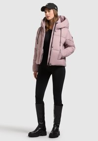 Svetloružová bundová páperová bunda s kapucňou, so zipsom a sťahovacími šnúrkami, v kombinácii s čiernymi legínami a čiernymi vysokými čižmami po kolená.
