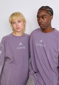 Chemises à manches longues violettes avec col rond, arborant le logo "adidas" et le texte "+TEAMGEIST", en tissu texturé et à coupe décontractée.