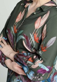Blusa verde con un motivo floreale in tonalità di rosa, rosso e blu. Presenta un colletto e maniche arricciate. Tessuto liscio e dalla consistenza. 