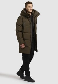 Olíva zöld puffernadrág, levehető kapucnival, cipzáras zsebekkel és steppelt mintázattal. Fekete nadrággal és csizmával viselve.