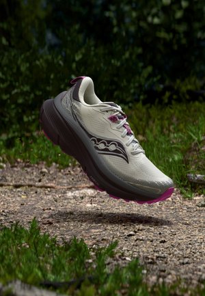 Scarpa da corsa con tomaia bianca, logo nero e suola nera spessa con dettagli rosa che fluttua sopra un sentiero di ghiaia e erba verde.