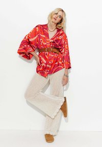 Haut de style kimono à motifs rouges et orange avec des manches larges, associé à des pantalons évasés texturés crème. Ceinture marron et chaussures assorties.