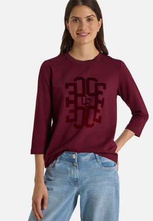 Femme portant un haut bordeaux à manches longues avec un motif géométrique sombre et un jean bleu clair, debout avec une main dans la poche.