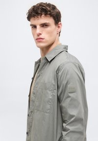 Jeune homme aux cheveux courts et bouclés portant une chemise gris clair à boutons avec une poche poitrine et le logo BOSS sur la manche.