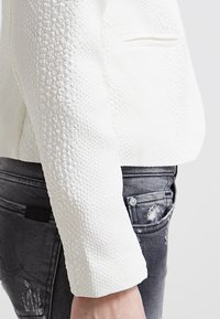 Veste blanche texturée à coupe ajustée, dotée d'un col rond et d'un motif subtil. Associée à un jean denim gris déchiré.