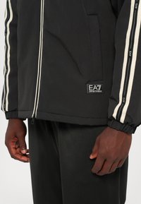 Črna jopa z zadrgo z belimi črtami po rokavih, elastičnimi manšetami in nalepko z logotipom EA7 na spodnji levi strani. Gladek, lahek material.