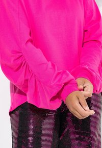 Sudadera rosa brillante con mangas recogidas, combinada con pantalones negros con lentejuelas. La tela parece suave con una textura lisa y acentos brillantes.