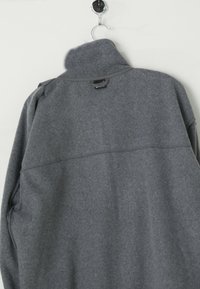 Graue Fleecejacke mit hohem Kragen und horizontalen Nahtdetails. Weiche Textur und lange Ärmel. Etikett in der Nähe des Kragens sichtbar. Hängt an einem Haken.