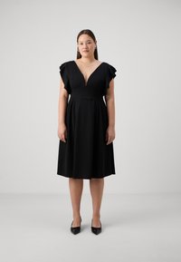 Vestido negro con escote en V profundo y mangas con volantes. Confeccionado en una tela suave, presenta un cuerpo ajustado y una falda acampanada. Combinado con tacones negros.