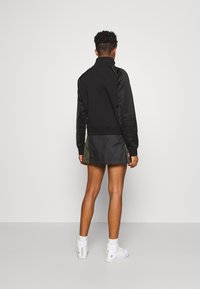 Svart sportig jacka med texturerade ärmar, kombinerad med en grön kjol. Vita högklackade sneakers fulländar looken. Enkel, funktionell design.