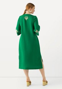Grünes Leinenkleid mit Dreiviertelärmeln, seitlichen Schlitzen und ausgeklügelter goldener Stickerei auf dem Rücken und an den Manschetten.