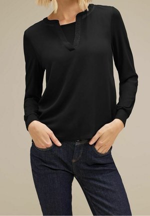 Kvinde iført en sort V-hals bluse med lange ærmer og satin kant, hænderne i lommerne, iført mørkeblå jeans mod en neutral baggrund.