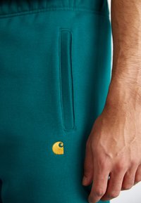 Teal Jogginghosen aus weichem Material mit einem kleinen goldenen Logo, sichtbarem Seitentasche und elastischem Bund für zusätzlichen Komfort.