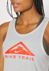 Hellgraues, ärmelloses Nike Trail Tanktop mit rotem dreieckigem Logo und dem Text "NIKE TRAIL." Atmungsaktiver Mesh-Stoff, sportliche Passform.