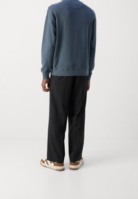 Blauwe katoenen sweatshirt met geribbelde mouwen, gecombineerd met zwarte plooibroeken en bruine en witte sandalen. De achteraanzicht laat een ontspannen pasvorm zien.