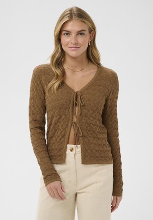 Jeune femme aux longs cheveux blonds portant un cardigan marron texturé noué devant et un pantalon crème taille haute, debout devant un fond blanc.