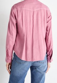 Chemise rose à manches longues avec un ourlet arrondi et des poignets boutonnés, confectionnée dans un tissu doux, avec un empiècement au dos. Portée avec un jean bleu.