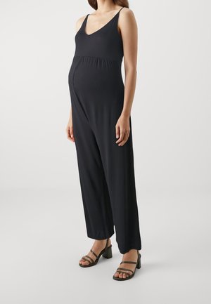 Schwarzer, ärmelloser Jumpsuit aus weichem Material mit V-Ausschnitt und elastischem Bund. Getragen mit schwarzen Sandalen mit Blockabsatz und Riemchen.