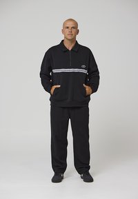 Jersey negro con media cremallera, con una franja horizontal blanca y logo; combinado con pantalones negros de corte recto y zapatillas negras.