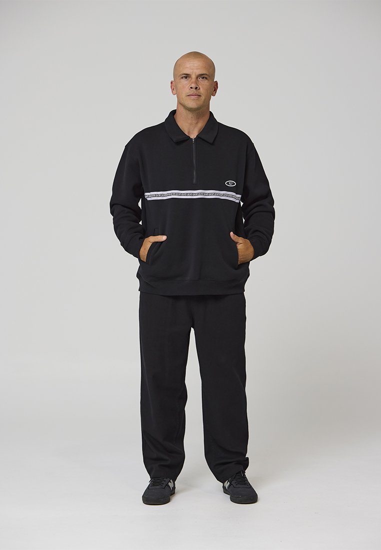 Jersey negro con media cremallera, con una franja horizontal blanca y logo; combinado con pantalones negros de corte recto y zapatillas negras.